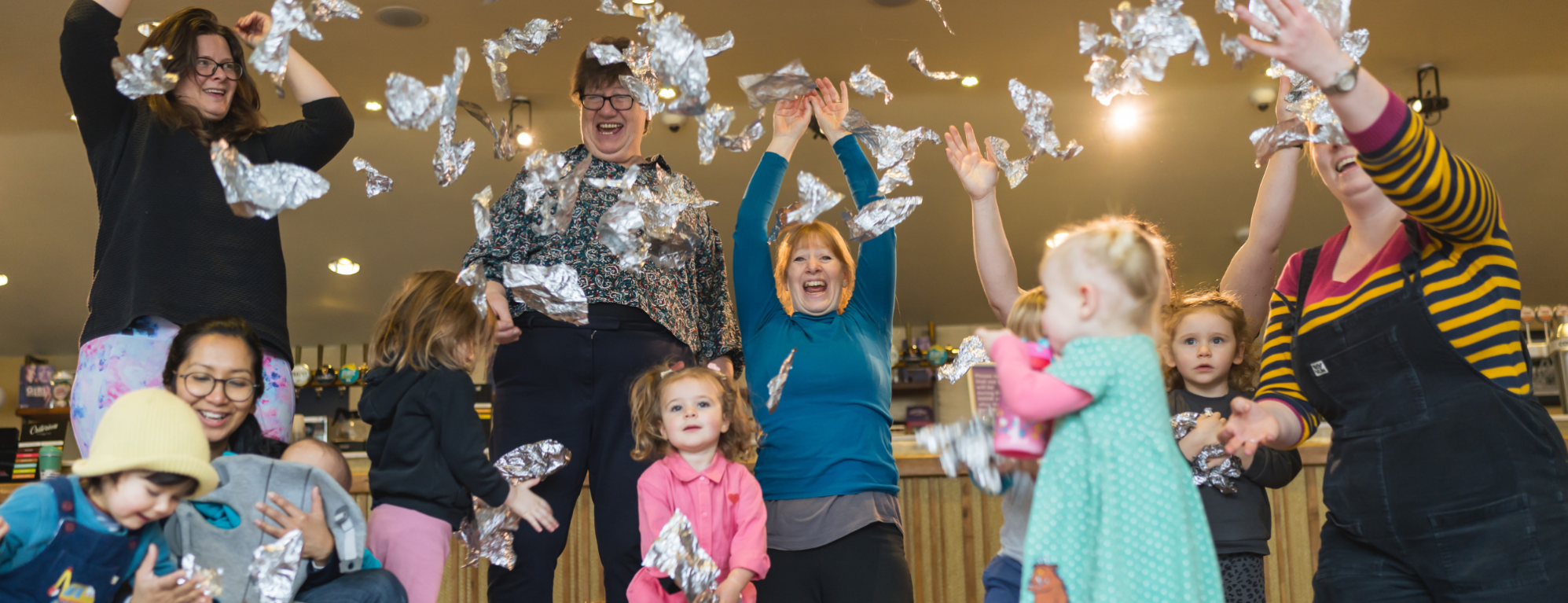 A group of adults and toddlers are standing in colourful clothes and throwing ripped up pieces of shiny silver tin foil in the air, smiling and laughing.