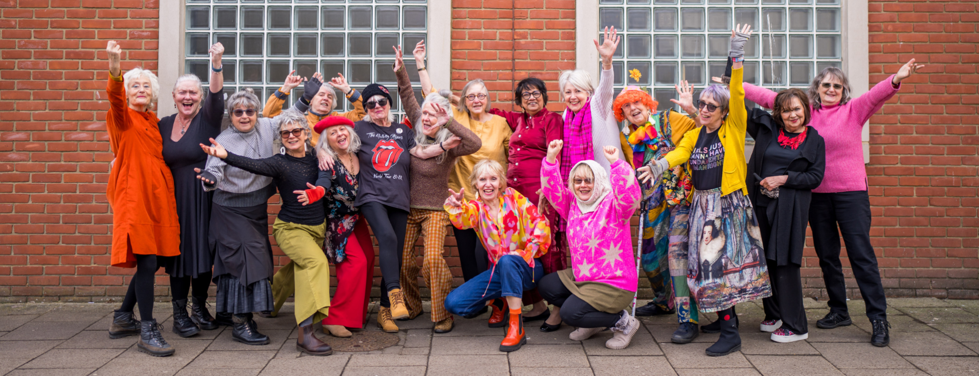 A large group of older individuals stand in front of a red brick wall. They are wearing vibrant colours like orange and pink, some our crouched. They are all posed looking at the camera, many with their arms up in the air and all smiling or laughing.