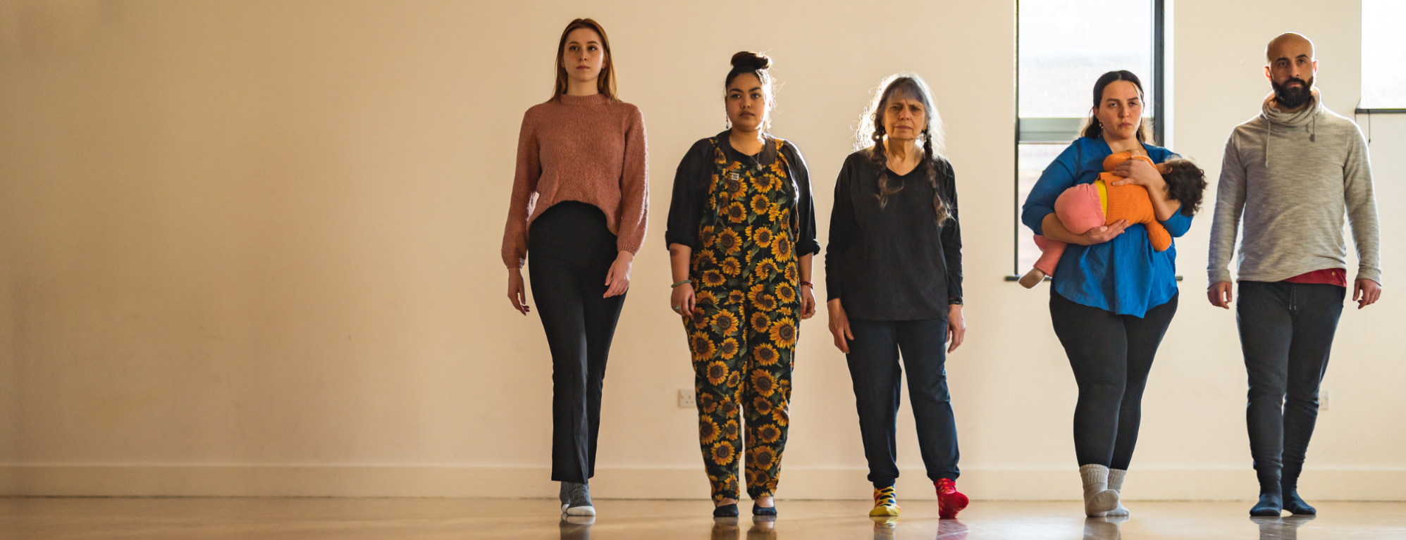 A group of five adults, one holding a small toddler to her chest, walk side by side in a line to a point beyond the camera. They are in a theatre studio space.